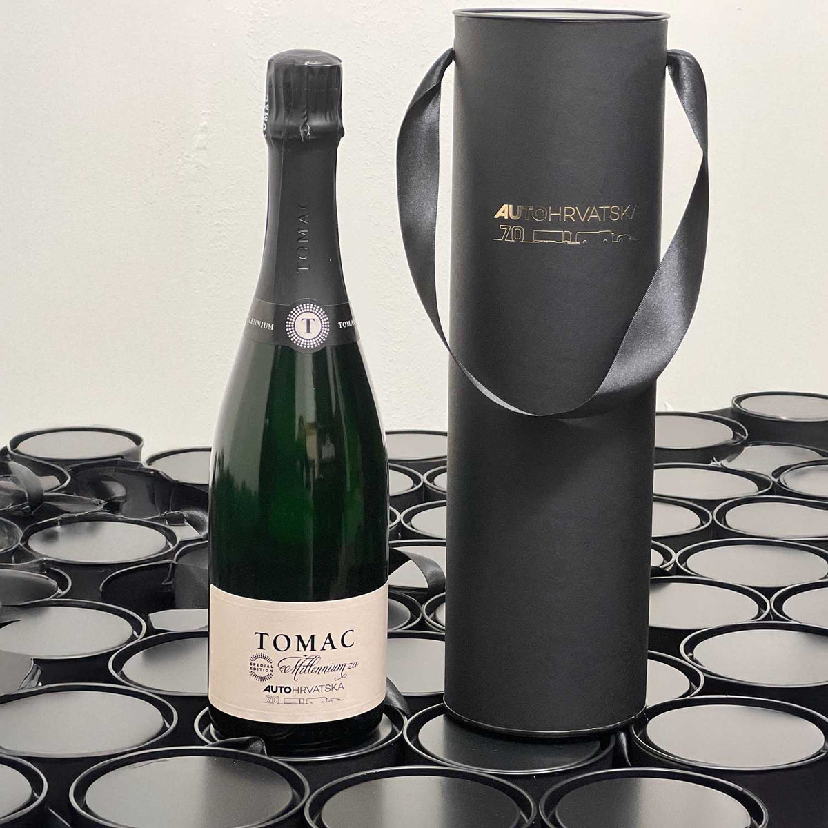 Tomac sparkling wine beside a black cylindrical gift box with gold Auto Hrvatska 70th-anniversary branding.