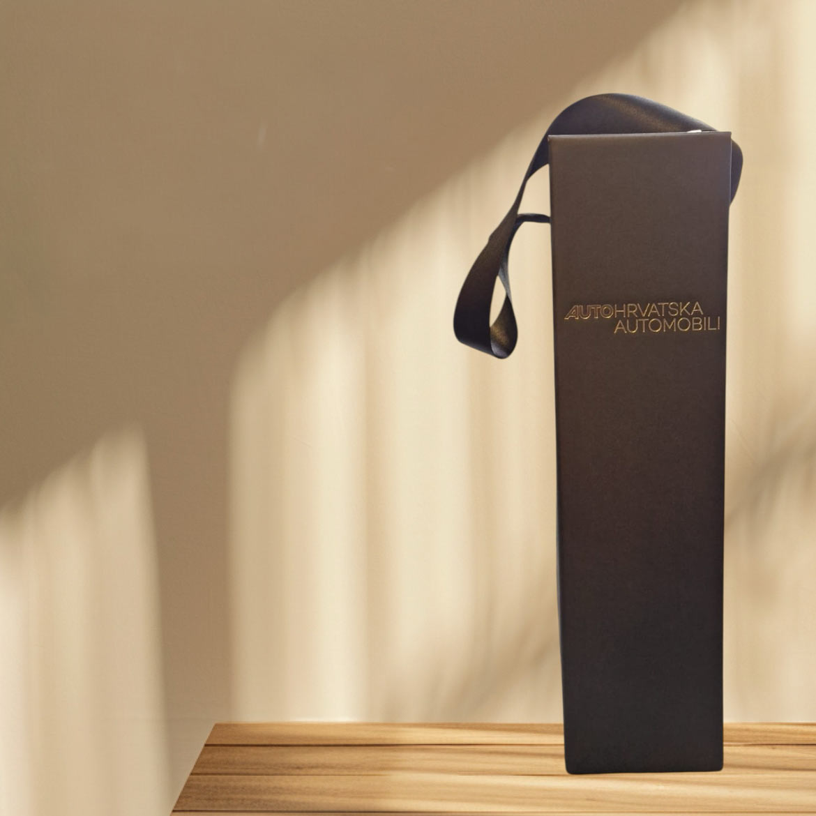 Black Auto Hrvatska Automobili gift packaging with gold branding and a satin handle, photographed in warm sunlight.